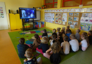 Grupa dzieci siedzi przed ekranem mobilnym, ogląda spektakl muzyczny.